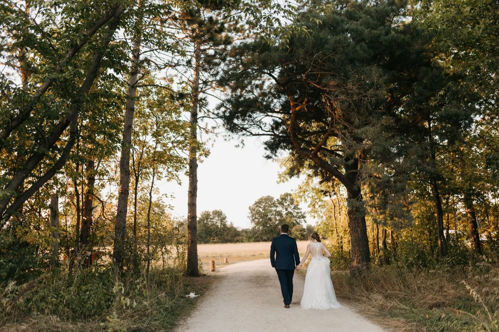 Bride and groom walk hand-in-hand down a tree-lined path at golden hour during their wedding day.