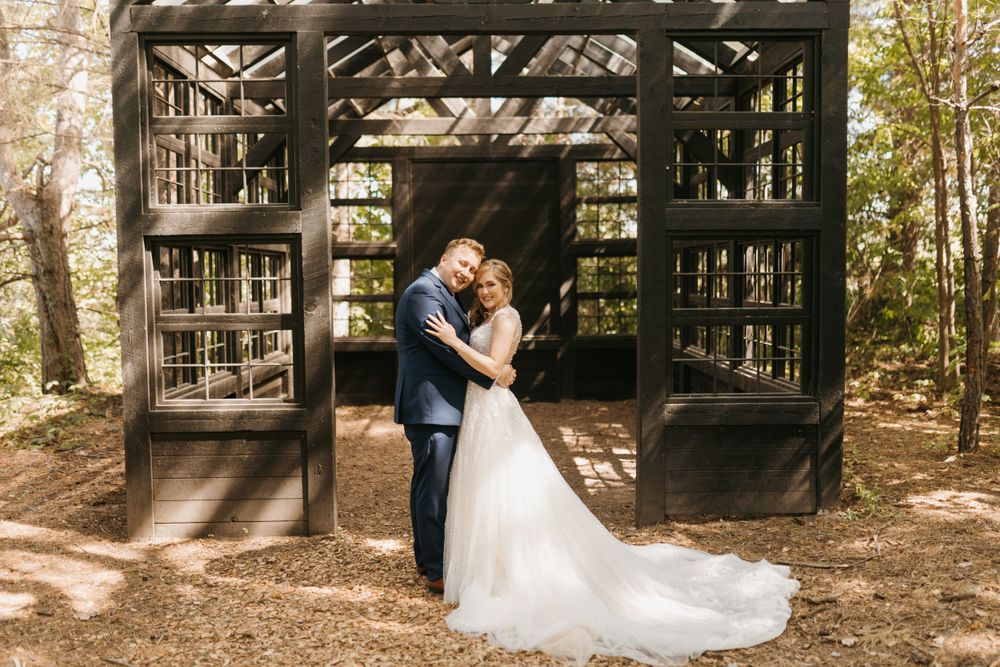 Bride and groom embrace lovingly in front of a rustic dark wooden greenhouse structure in a wooded setting.
