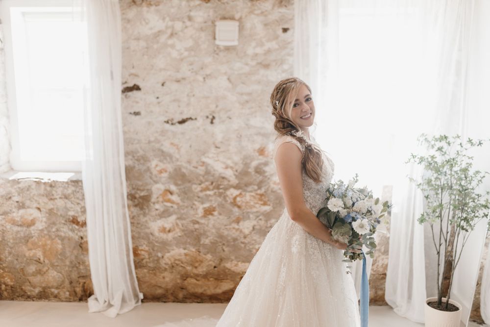 Bride in lace A-line wedding gown smiles over shoulder while holding blue floral bouquet in bright rustic venue.