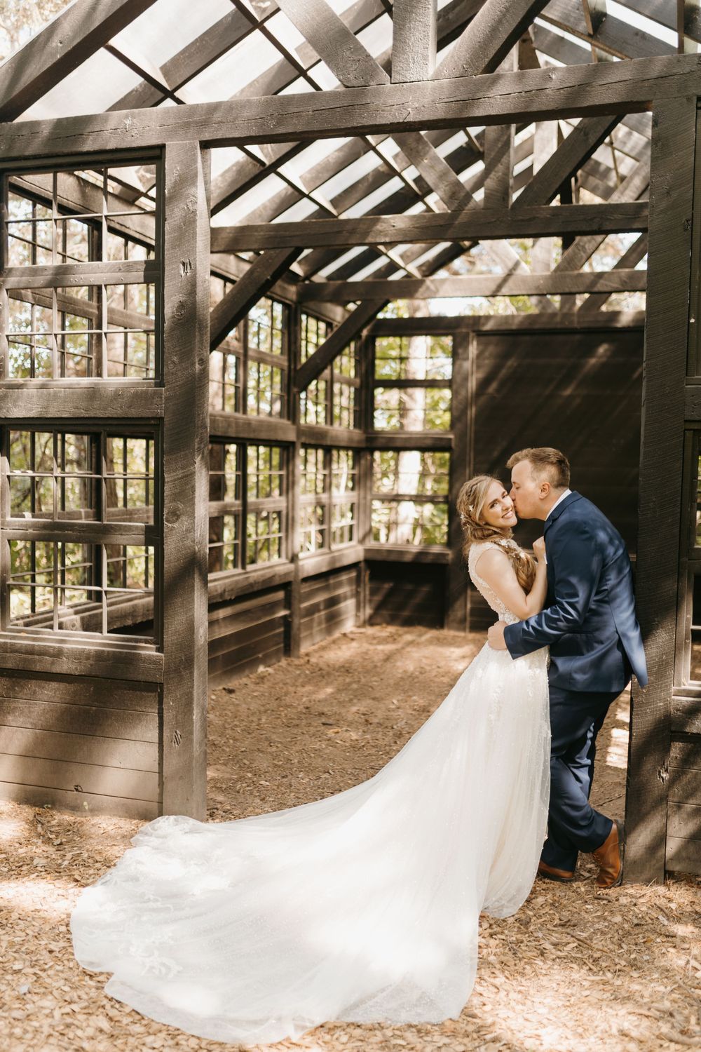 Bride in flowing white gown and groom in navy suit kiss inside a wooden greenhouse.