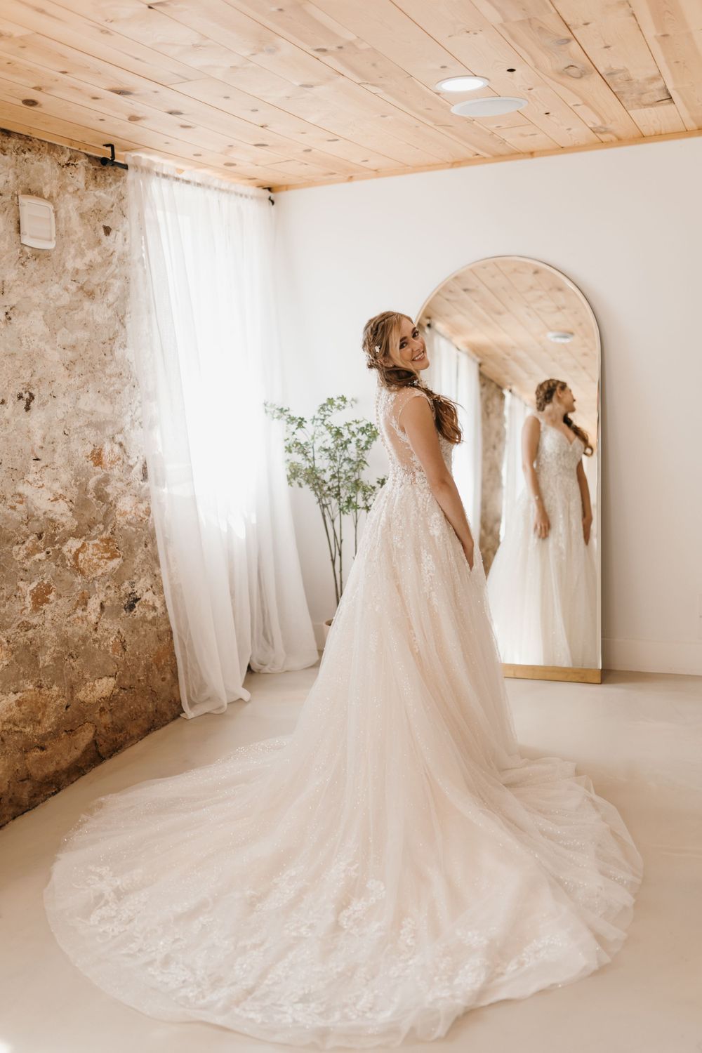 Bride in elegant lace A-line wedding gown with long train poses beside arched mirror in bright rustic bridal suite.