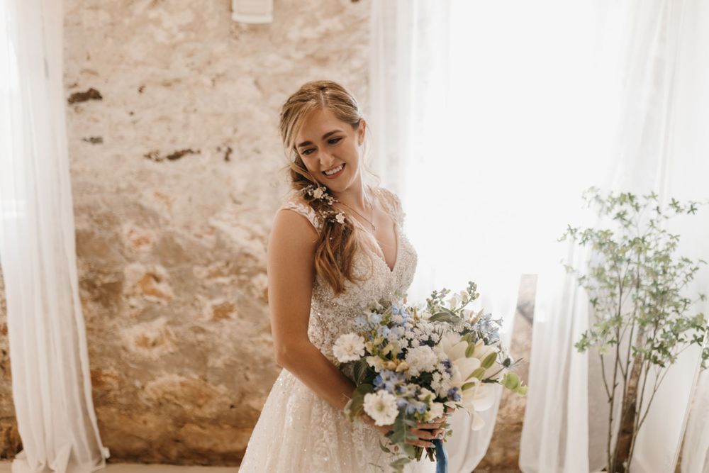 Glowing bride in lace wedding dress smiles while holding a blue and white floral bouquet in a rustic sunlit room.