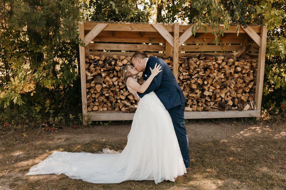 Bride and groom share a dip kiss in front of a rustic outdoor firewood storage rack.