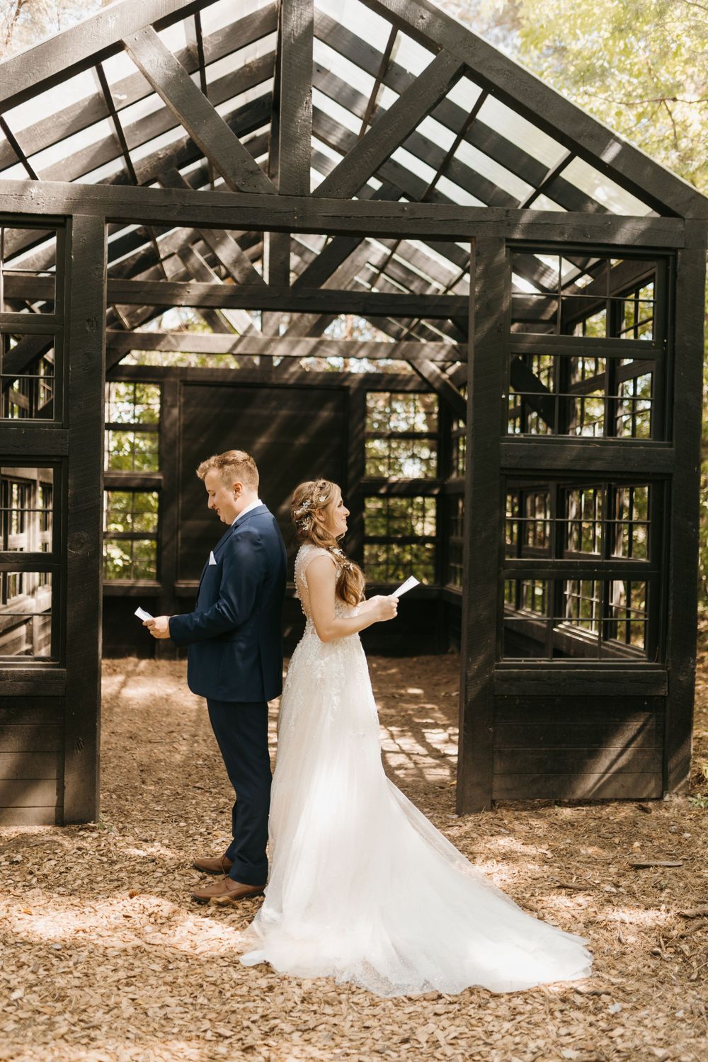 Bride and groom stand back-to-back reading vows outside a rustic dark wooden greenhouse structure in a forest.