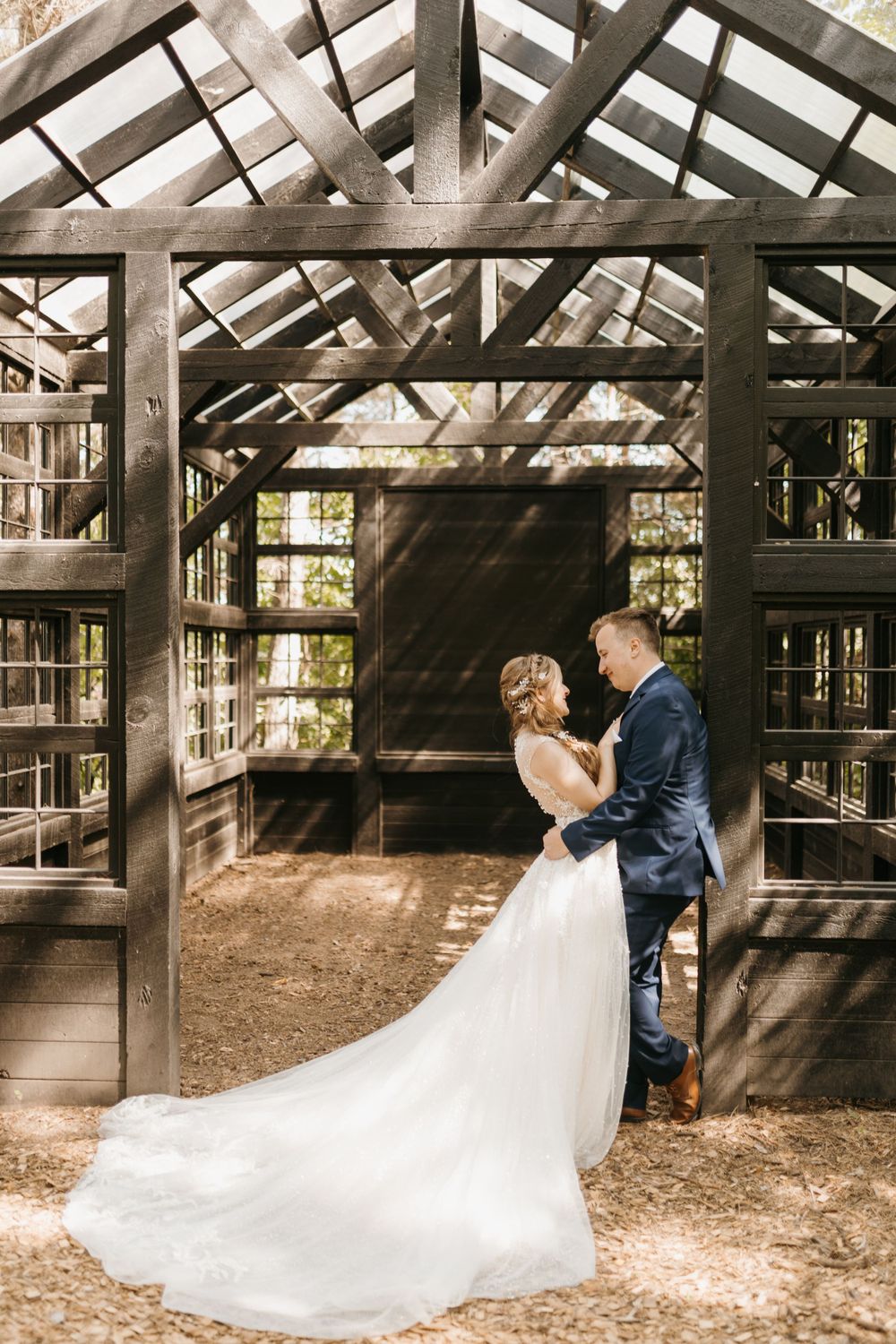 Bride and groom embrace tenderly inside a dark wood greenhouse chapel in a forest setting.