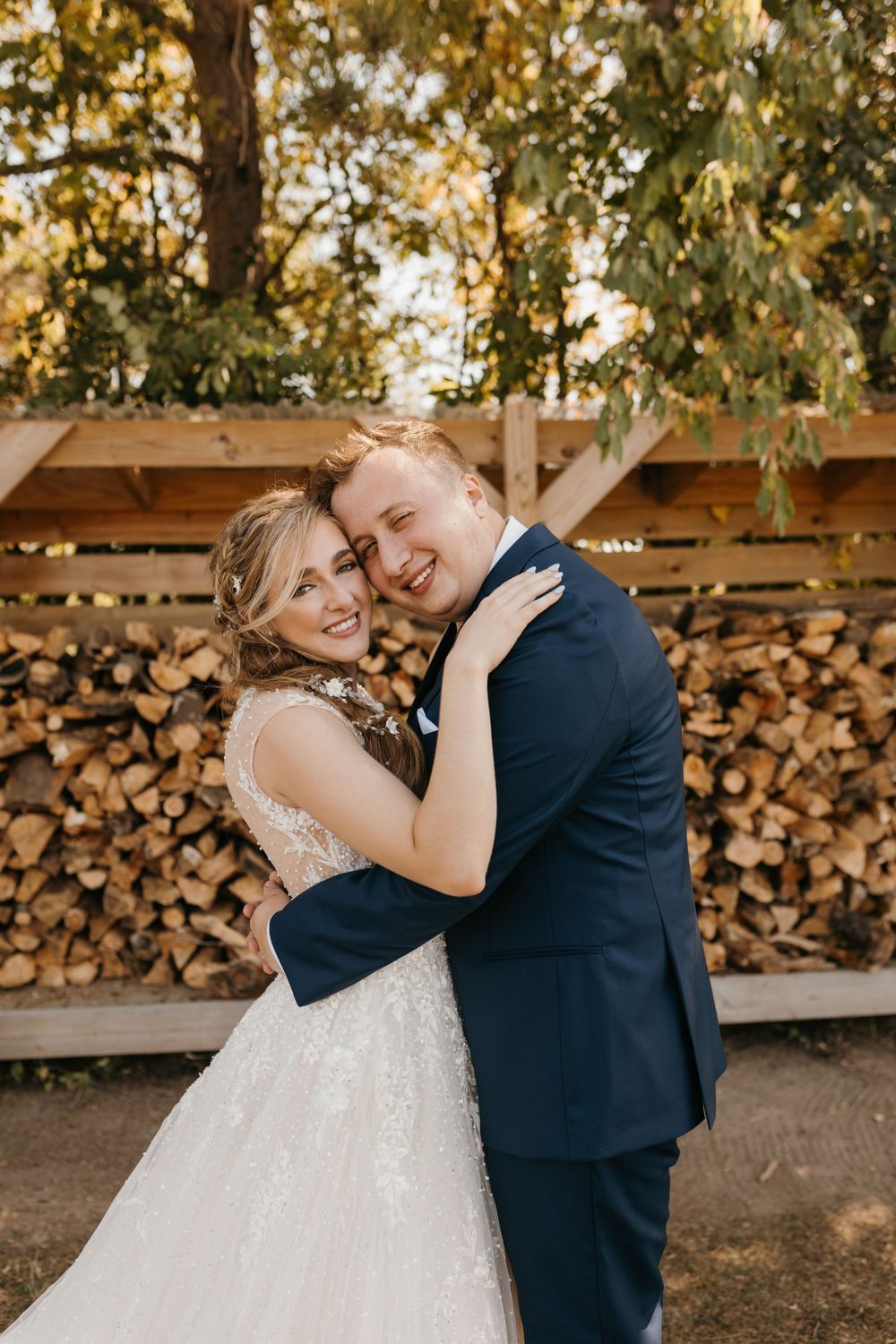 Smiling bride and groom embrace joyfully in front of a stacked firewood rack outdoors.