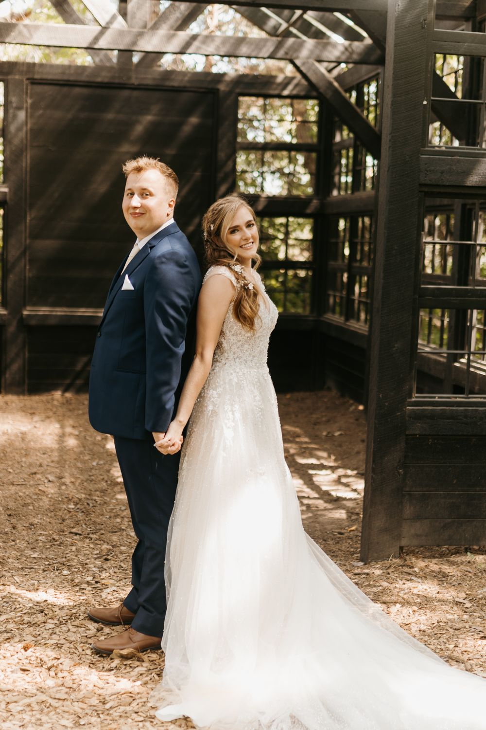 Bride and groom hold hands back-to-back smiling at camera outside a dark wooden greenhouse wedding venue.