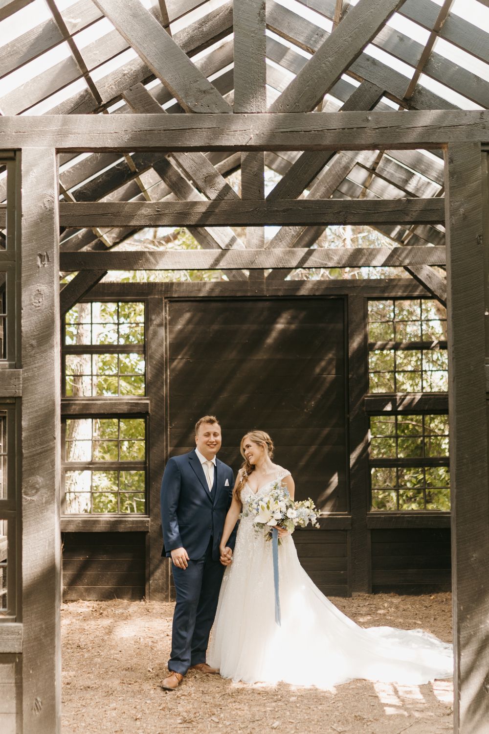 Bride holding bouquet and groom pose inside a rustic open-roof wooden greenhouse wedding venue in the forest.