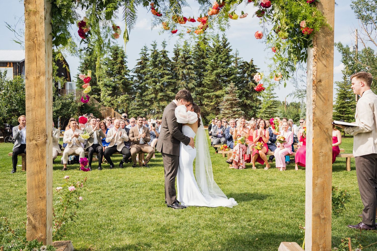 Outdoor wedding ceremony under wooden arch decorated with flowers and greenery.