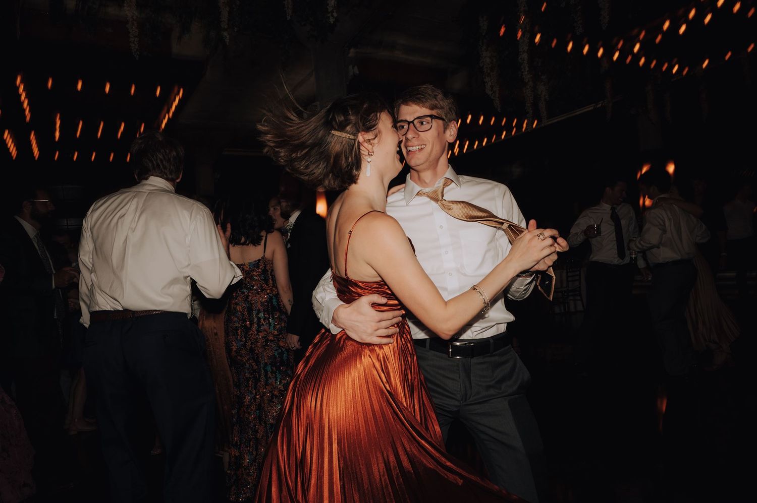Wedding guests dancing under string lights at a dimly lit reception venue.