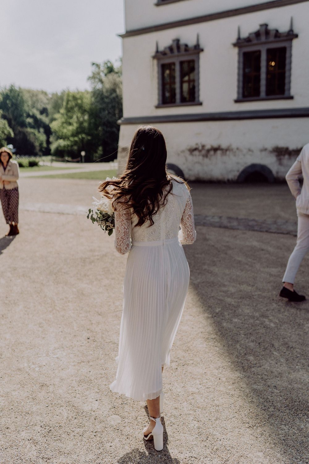 Bride in pleated white dress walks away from vintage building on gravel path.