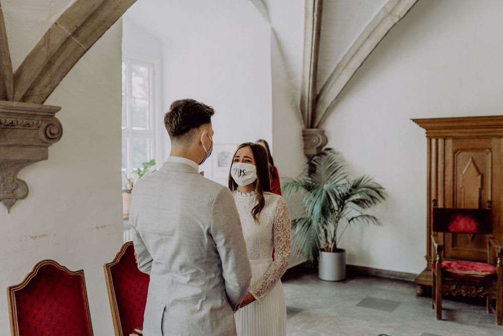 A couple wearing protective face masks stands together in an elegant room with arched ceilings and red velvet chairs.