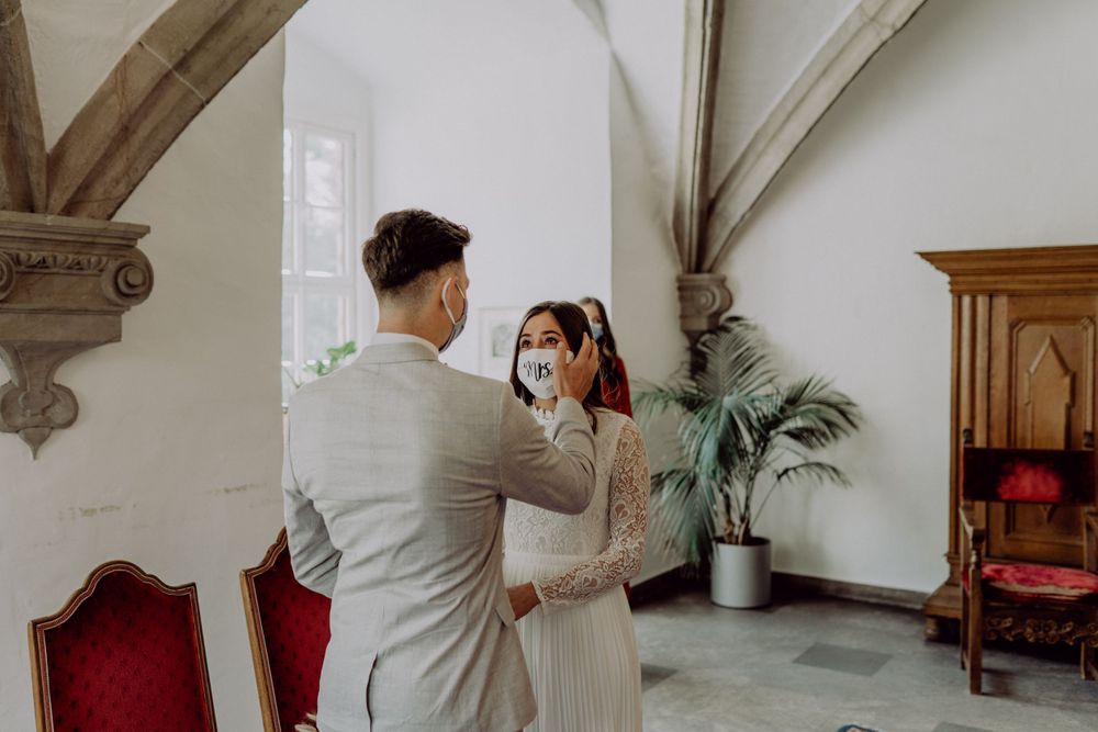 A couple in formal attire and protective masks sharing a tender moment in front of ornate architectural details.
