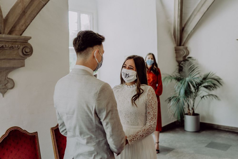 A couple exchanges vows in an elegant room while an officiant in red dress observes from the background.