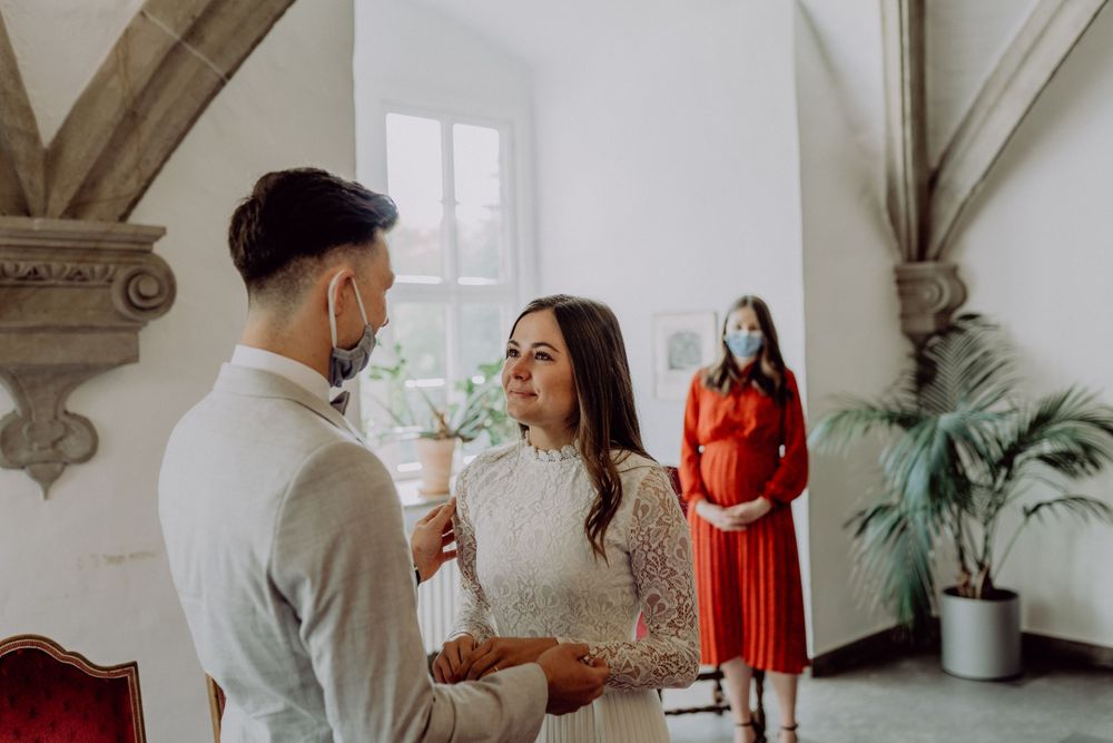 A couple shares an intimate moment in a white room with ornate architectural details while a woman in red watches.