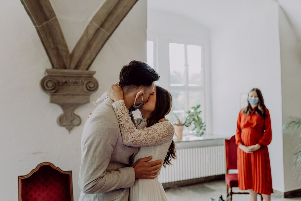 Intimate embrace between a couple in an elegant white interior while someone in red observes from afar.