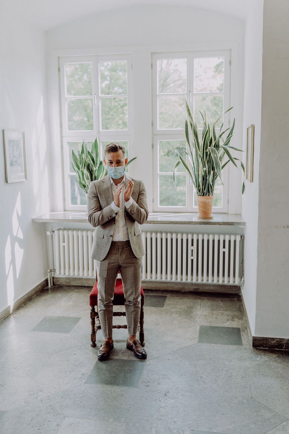 A person in a grey suit stands by a bright window with indoor plants and vintage radiator.