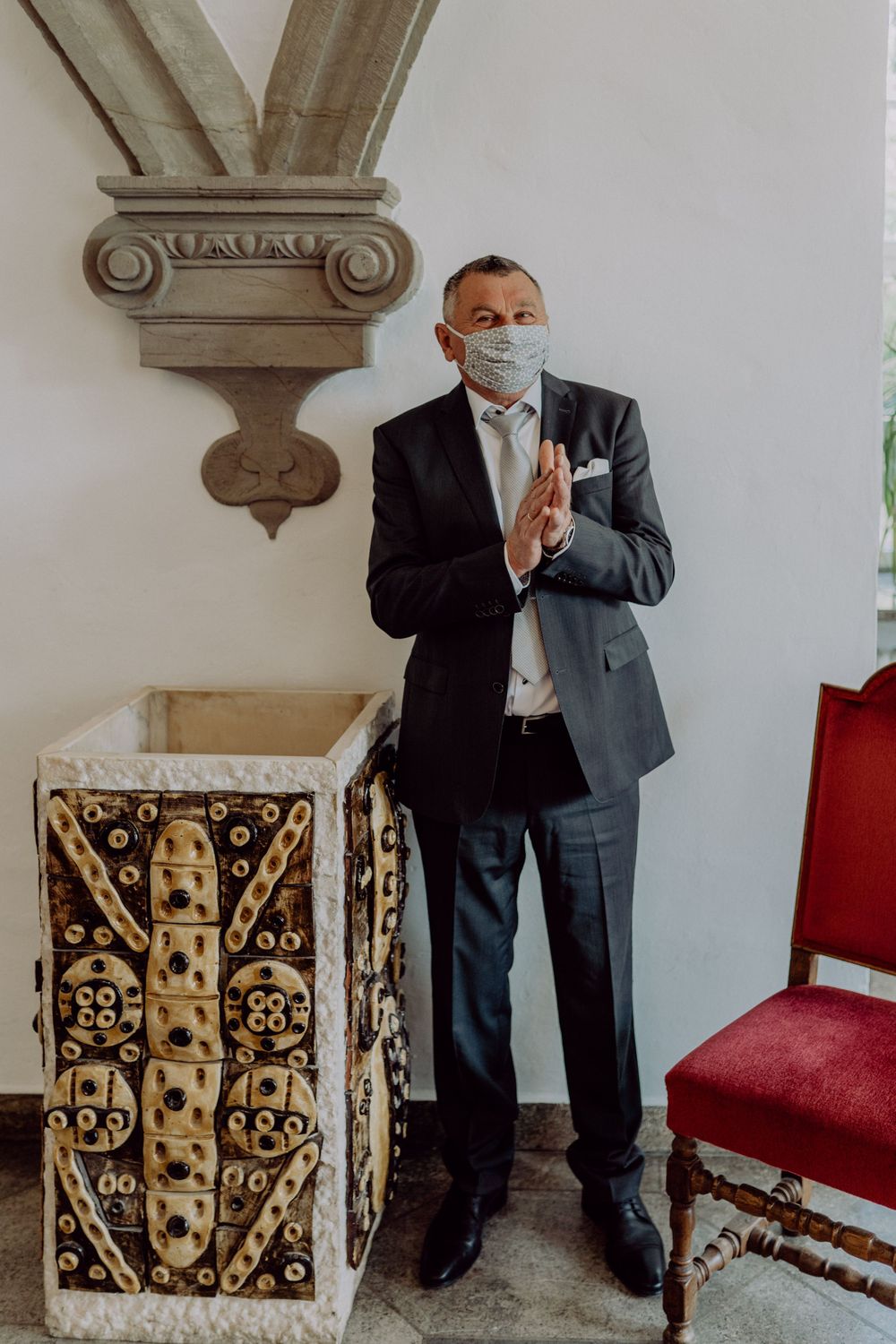A person in a dark suit and face mask stands praying next to decorative architectural details.