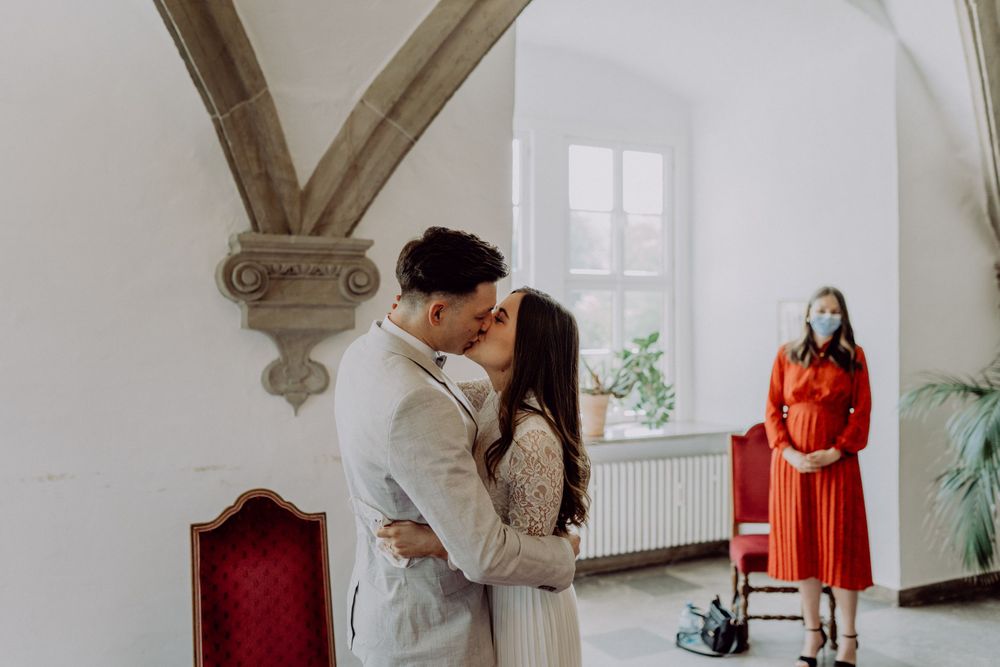 A couple embraces in a bright room with arched ceilings while an officiant in red looks on.