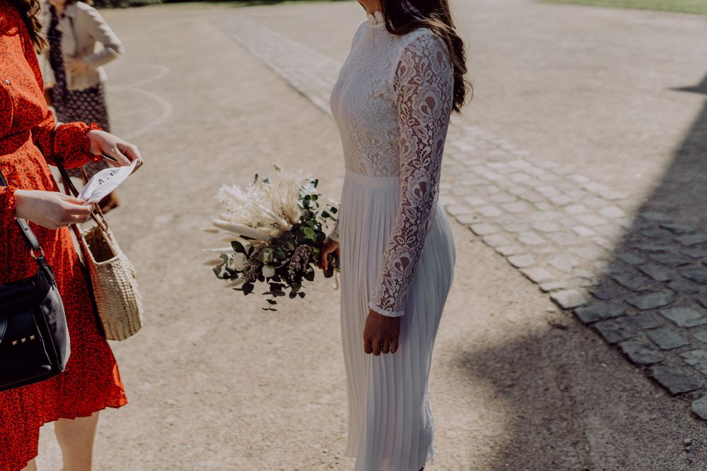 A person in a white lace wedding dress holds a delicate bouquet against a stone pathway.