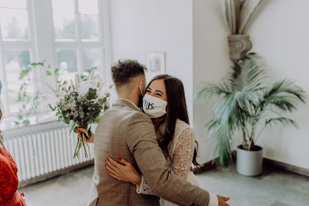 Couple wearing face masks embrace in a bright room with indoor plants and window light.