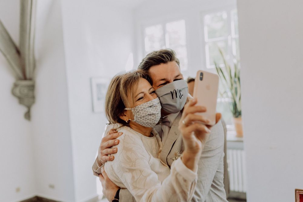 Couple wearing protective masks take a selfie while embracing in a bright interior space.