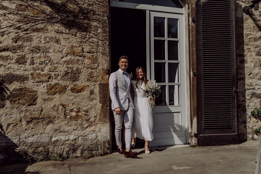 Newlyweds stand together in doorway of historic stone building with wooden shutters.