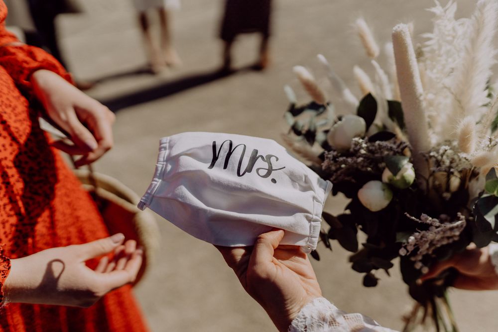 Close-up of hands holding a white face mask with 'Mrs' text during a wedding ceremony.