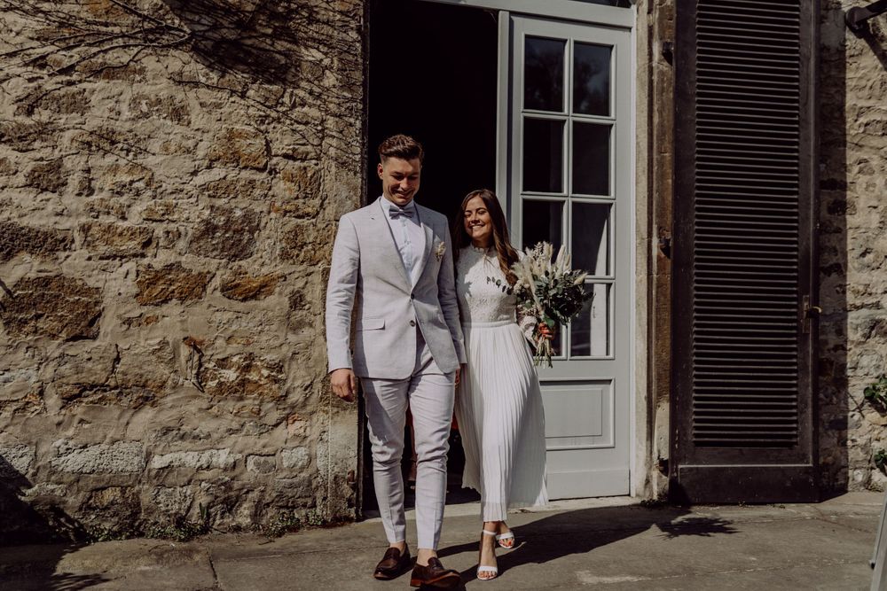 Wedding couple poses in formal attire against stone wall with white door and shutters.