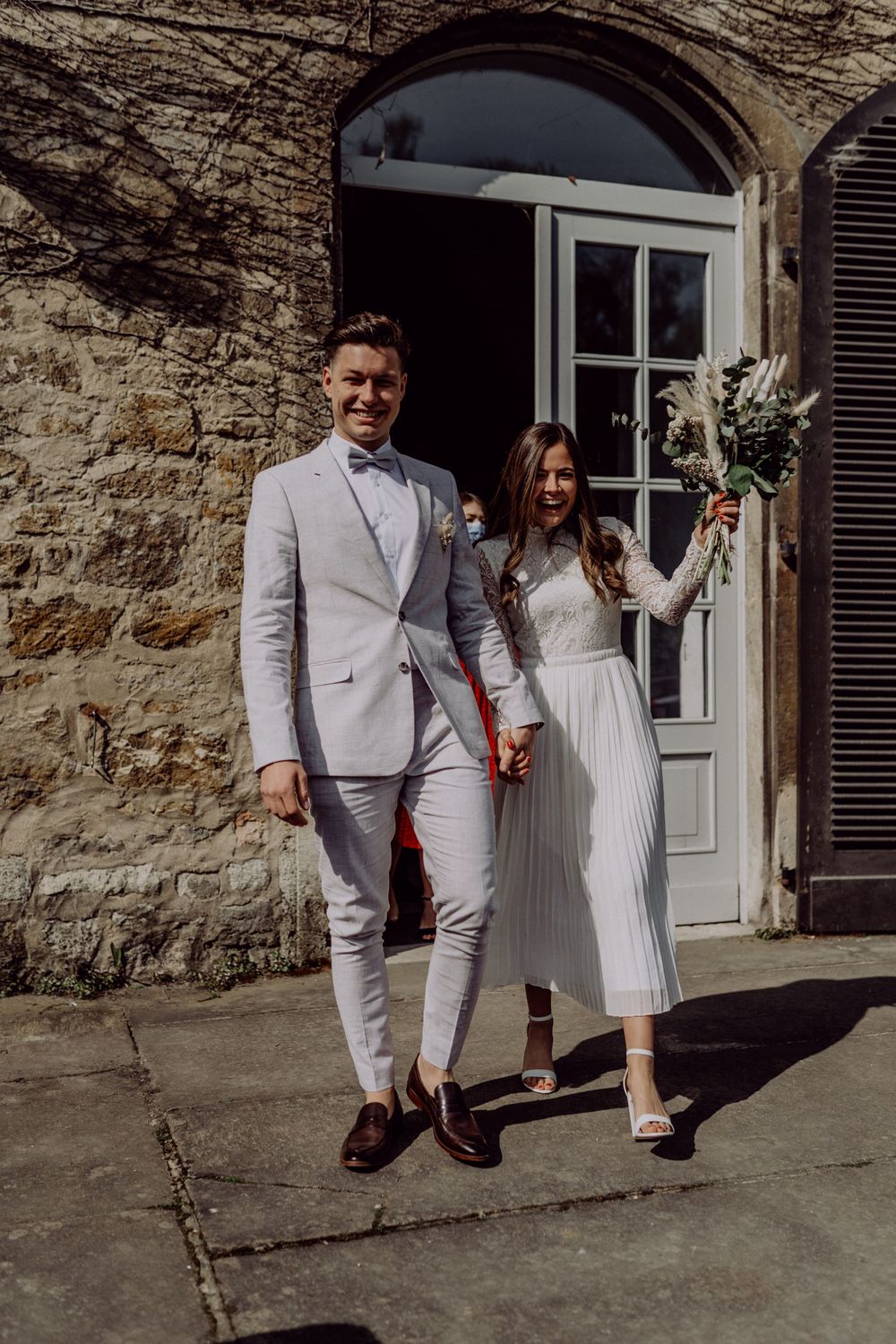 Elegant couple in light-colored formal wear pose outside historic stone building entrance.
