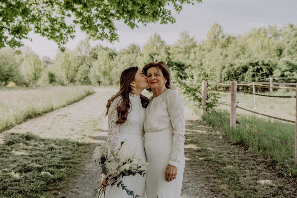Two people in white outfits share an intimate moment in a sunlit garden with trees and a wooden fence.