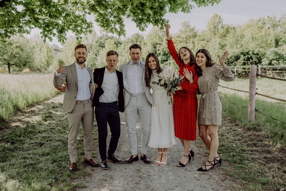 A wedding party poses together outdoors with guests in formal attire against a natural backdrop.