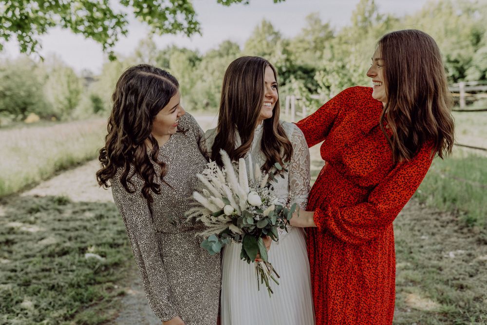 Three friends share joyful laughter while holding a white floral bouquet in an outdoor garden setting.
