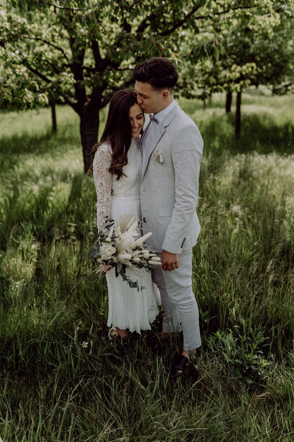 A romantic moment captured between newlyweds in white attire amidst tall grass and blooming trees.