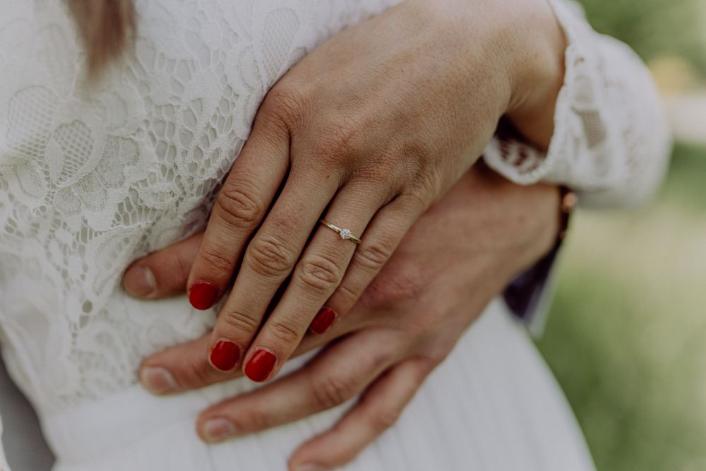 Close-up shot of hands with red nail polish and wedding ring against white lace fabric.