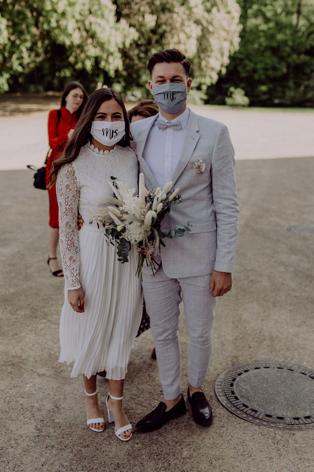 A couple in wedding attire wearing matching face masks pose with a bouquet outdoors.