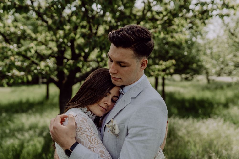 Tender embrace between couple in wedding attire surrounded by spring greenery.
