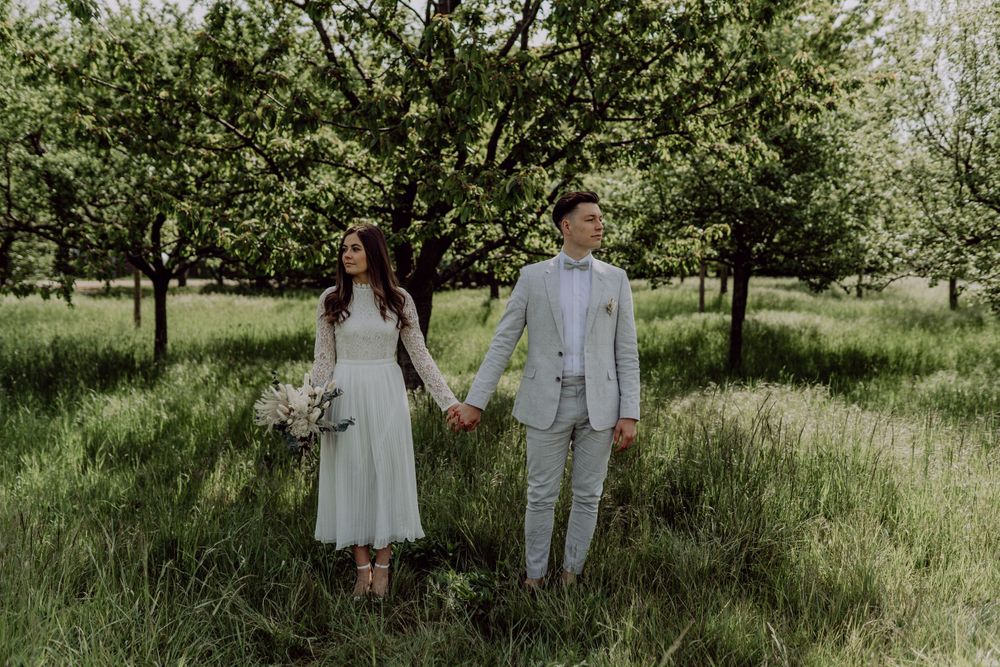 Wedding couple holding hands in orchard wearing light grey suit and white dress.