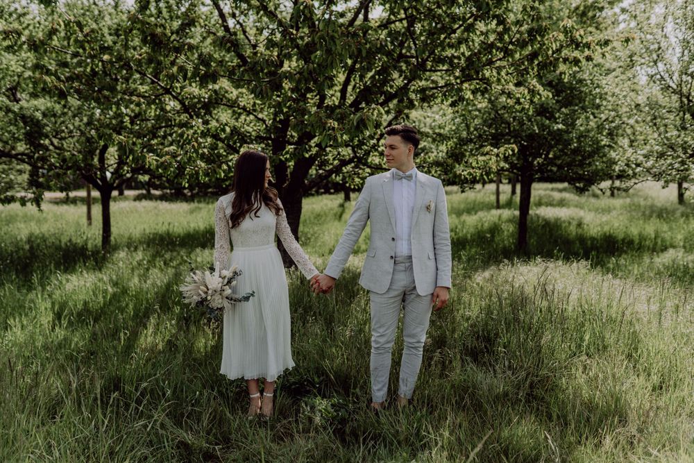 Full-length portrait of couple in wedding attire standing in spring orchard.