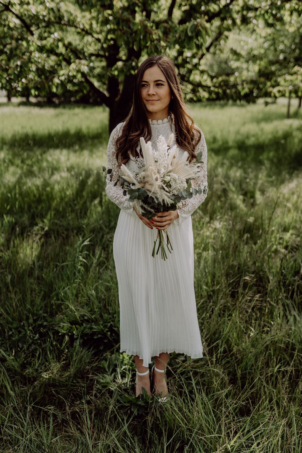 A white lace dress with long sleeves is worn in an outdoor garden setting with a bouquet of dried flowers and greenery.