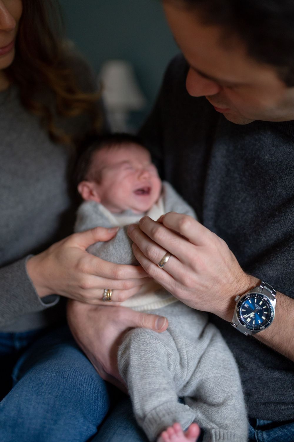 Laughing baby in grey outfit being held by both parents.