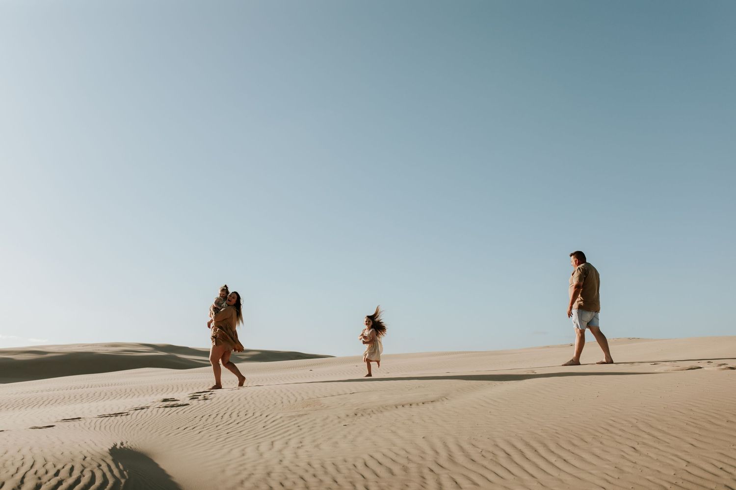 Three people walking across vast sand dunes under a clear blue sky.
