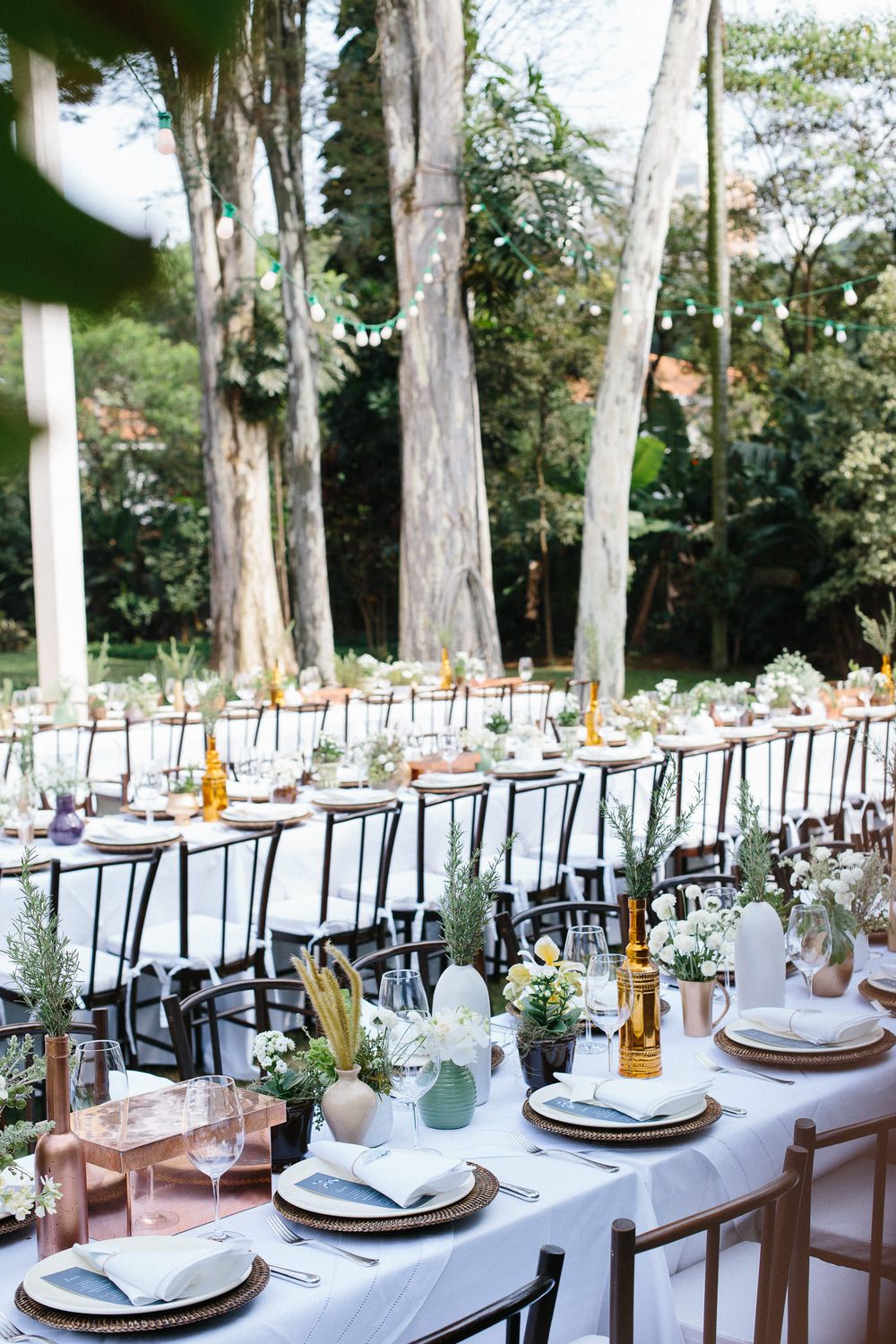 Outdoor wedding reception table setup with white linens and floral centerpieces among tall trees.