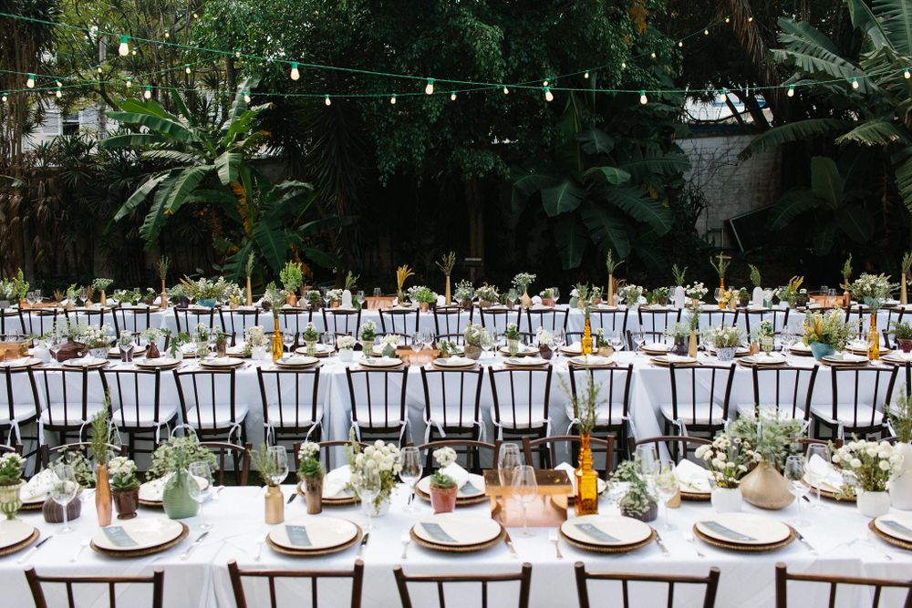 Long outdoor wedding reception table setup with string lights and lush greenery decorations.