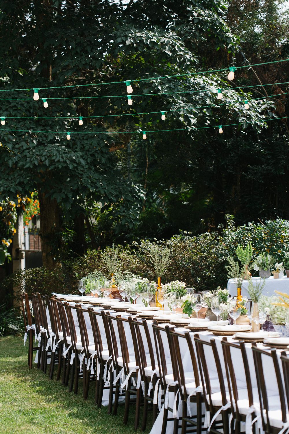 Garden reception table under string lights with white linens and dark wooden chairs.