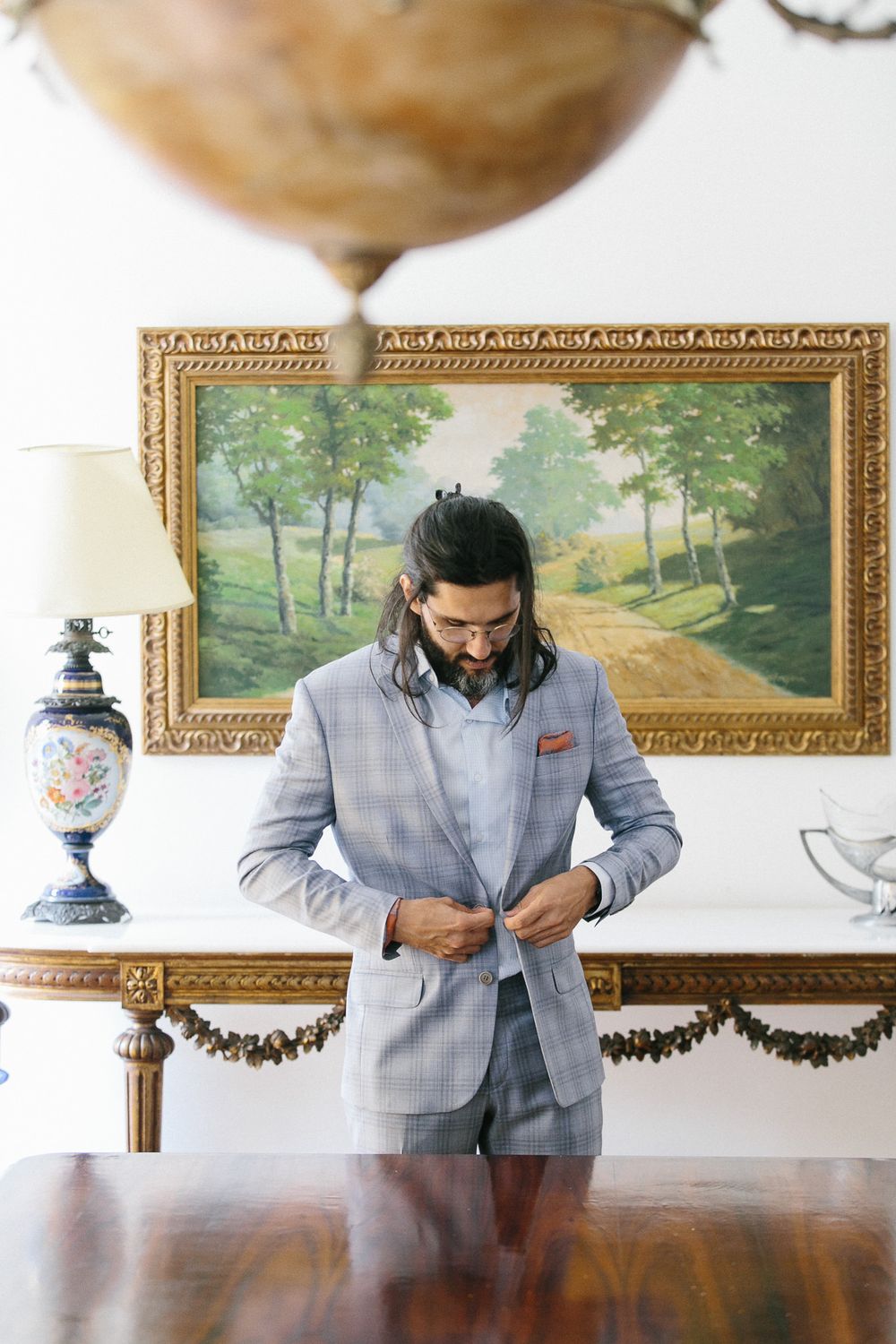 Person in light gray suit adjusting clothing in formal room with antique furniture and artwork.