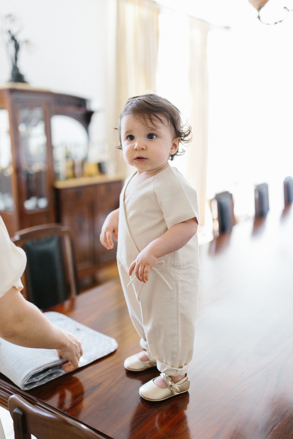 Small toddler in white outfit standing on polished wooden table.