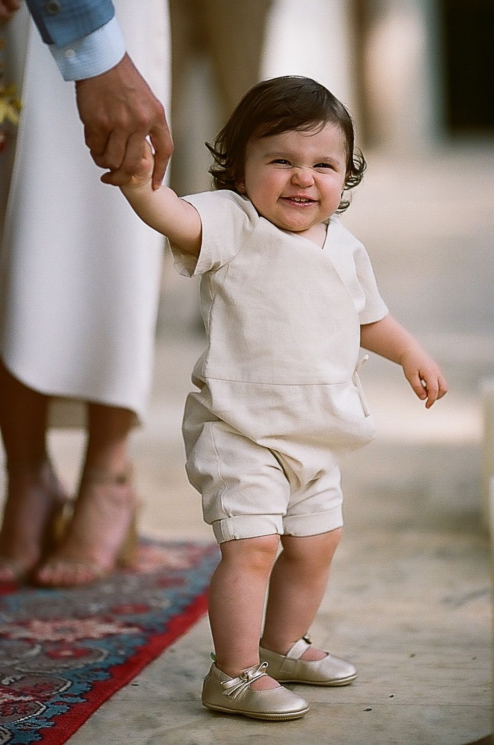 Toddler in white outfit taking first steps on a decorative rug.