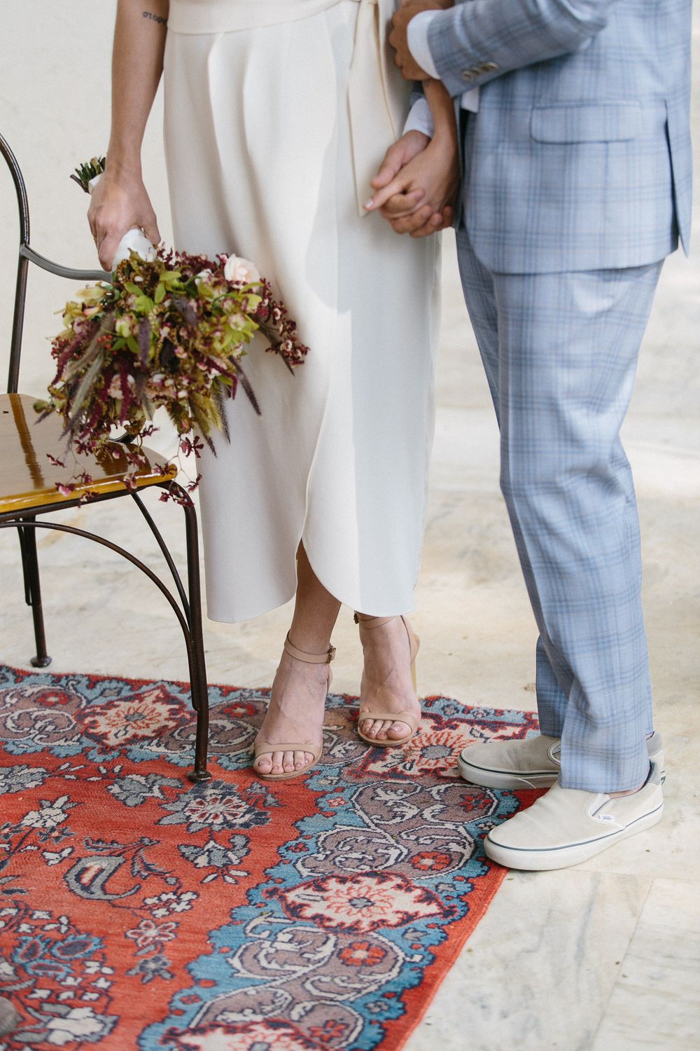 A couple holding hands in wedding attire stands on a decorative red carpet with a bouquet of dried flowers.