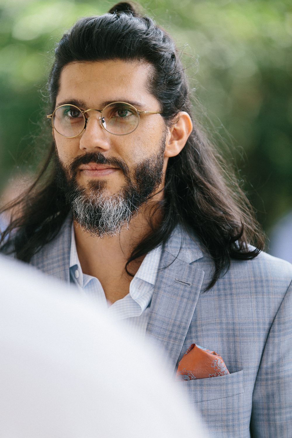 Portrait of a person in a light gray suit with long hair and glasses against a blurred background.